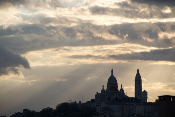 The image features a silhouette of the Basilica of the Sacred Heart in Paris under a dramatic sky with sunbeams piercing through the clouds.
