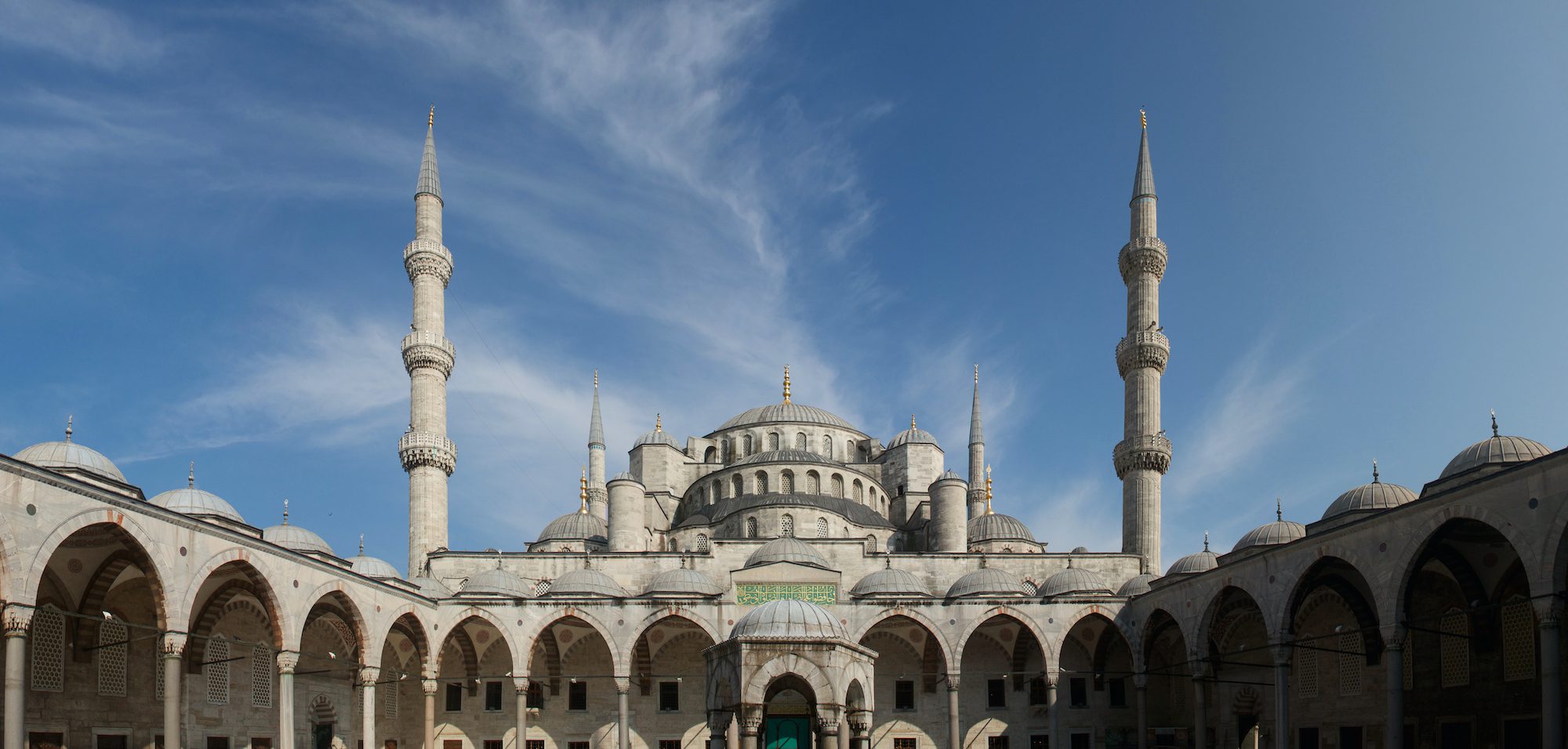 This is an image of a large, historical mosque with multiple domes and slender minarets against a clear blue sky, intricately designed with an open courtyard.