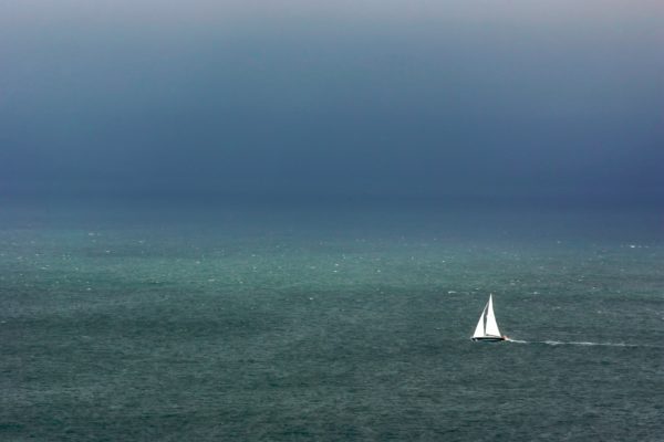 A lone sailboat with white sails navigates across a vast, dark blue sea under an overcast sky, creating a sense of solitude and tranquility.