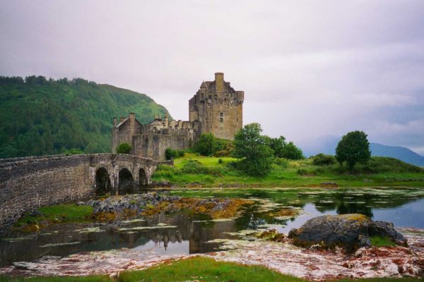 A historic castle with a stone bridge sits beside a calm body of water, surrounded by lush greenery and hills under a cloudy sky.