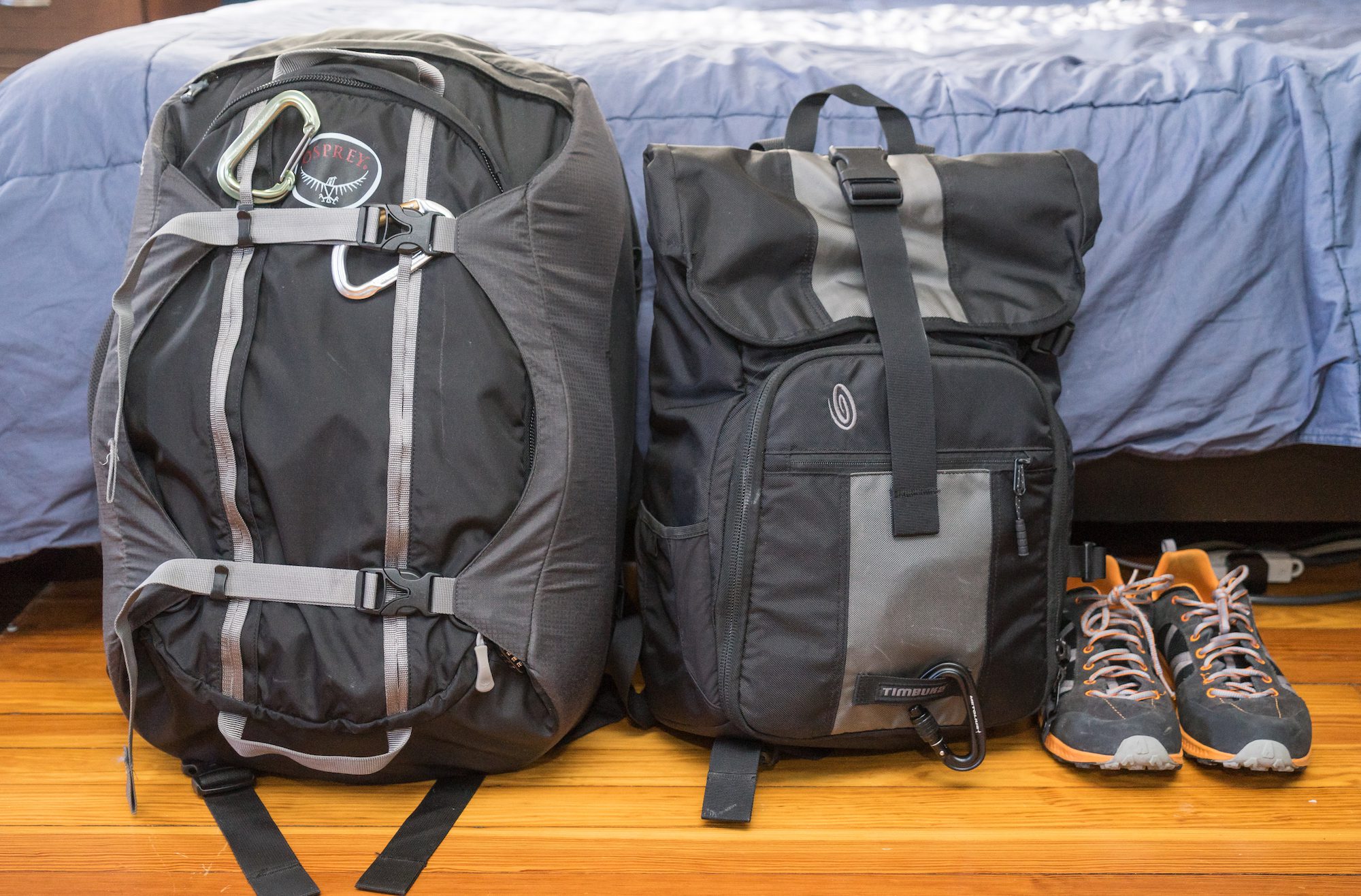 A large grey backpack with external straps and a smaller black bag are placed on a wooden floor, next to a pair of grey-orange sneakers, indoors.