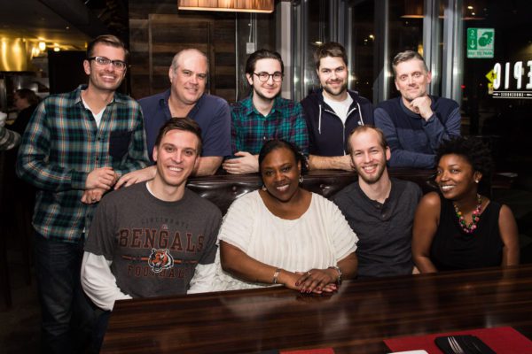 A diverse group of eight smiling adults sitting and standing around a restaurant table. Indoors, casual clothing, seems like a friendly gathering or celebration.