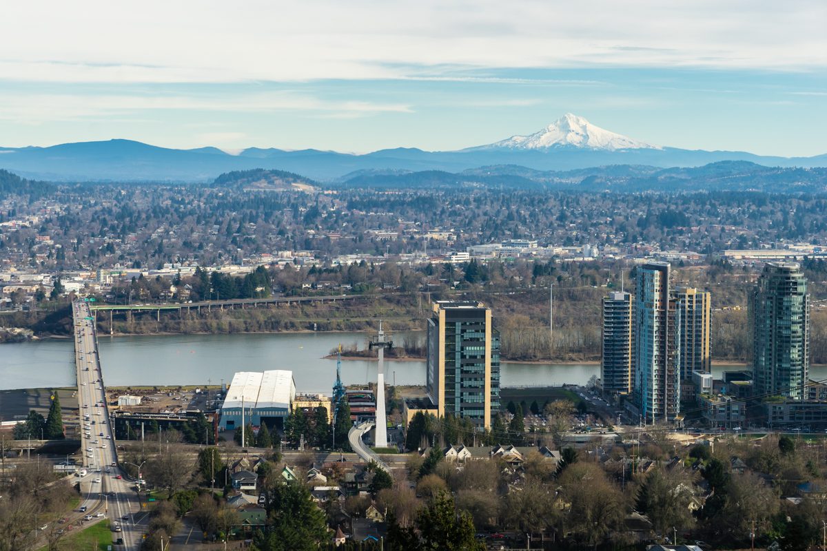 This image depicts a city skyline with high-rise buildings beside a river, a bridge under construction, and a snow-capped mountain in the background.