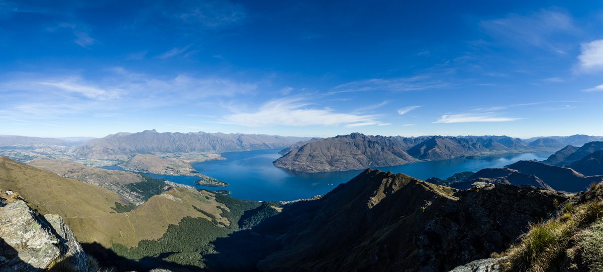 Panoramic view from a mountain peak showing a vast blue lake surrounded by rugged terrain and rolling hills under a clear sky.
