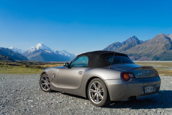 A silver convertible car (BMW Z4) is parked on a gravel surface with snow-capped mountains and a clear blue sky in the background.