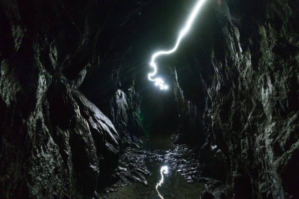 This image shows a dark, narrow cave illuminated by a serpentine trail of light, possibly from a headlamp, leading towards the distant, sunlit cave exit.