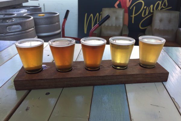 A flight of five different beers is presented in small glasses on a wooden paddle on a rustic table, showcasing a range of colors from pale yellow to amber.