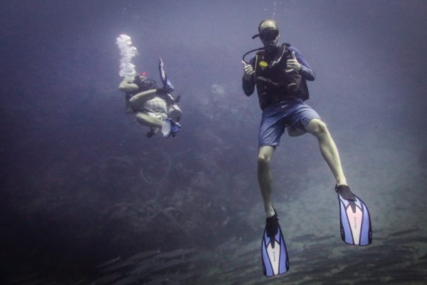 Two people are scuba diving underwater. The person in the foreground is wearing fins and holding diving equipment, while hazy blue water surrounds them.