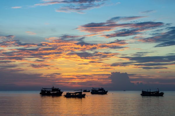 Several boats are silhouetted against a vibrant sunrise over a calm ocean, with clouds streaked in shades of orange, yellow, and blue.