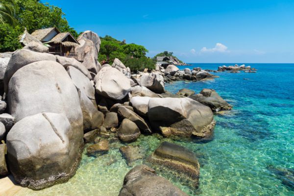 A serene tropical beach scene with clear turquoise water, large smooth boulders, lush greenery, and a wooden hut nestled among the rocks under a blue sky.