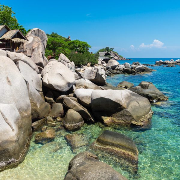 A serene tropical beach scene with clear turquoise water, large smooth boulders, lush greenery, and a wooden hut nestled among the rocks under a blue sky.