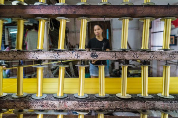 A person stands behind a traditional weaving apparatus with multiple spools of yellow threads. The focus is on the weaving machine in a workshop setting.