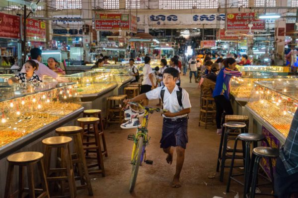 A young boy pushes a bicycle through an indoor market with rows of bright, illuminated jewelry counters while people browse and vendors attend stalls.