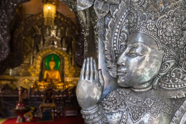 An intricately carved silver sculpture of a face with a hand raised is in the foreground, with a serene golden Buddha statue visible in the background.