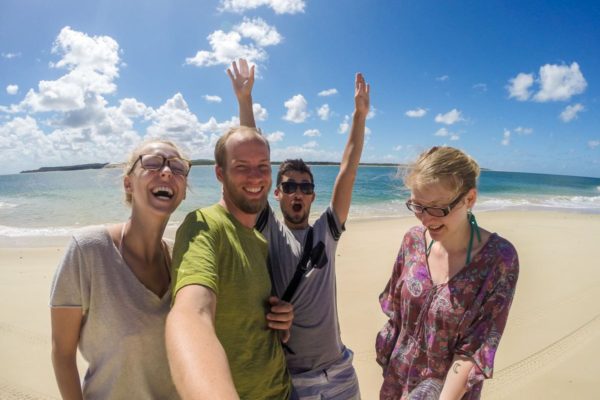 Four people are joyfully taking a selfie on a sunny beach with clear blue skies and the ocean in the background. They seem happy and relaxed.