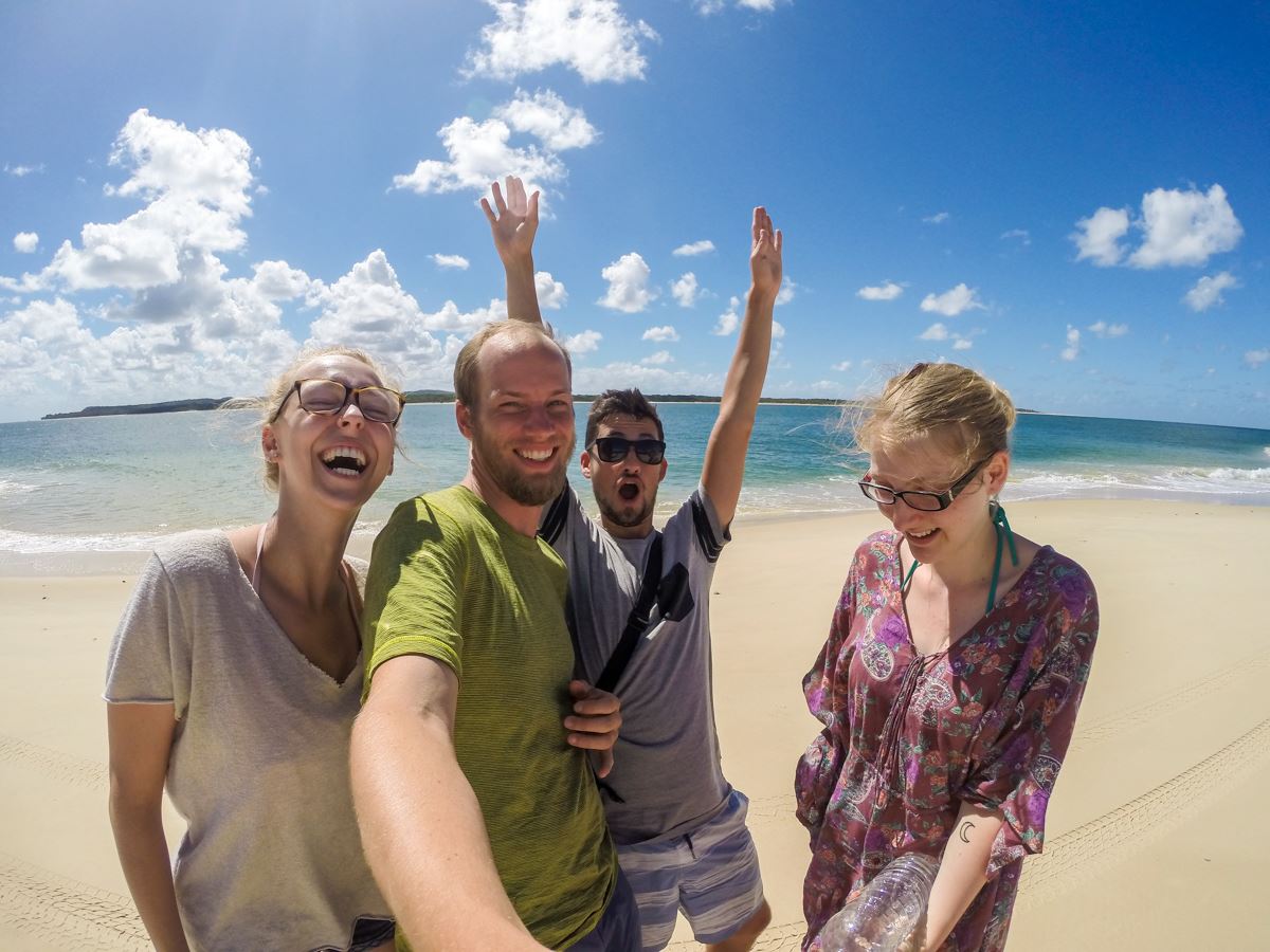 Four people are joyfully taking a selfie on a sunny beach with clear blue skies and the ocean in the background. They seem happy and relaxed.