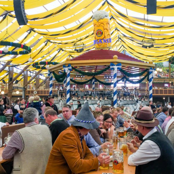 Inside a bustling festival tent with people wearing traditional attire seated at long tables, enjoying large steins of beer under vibrant yellow-and-white striped canopies.