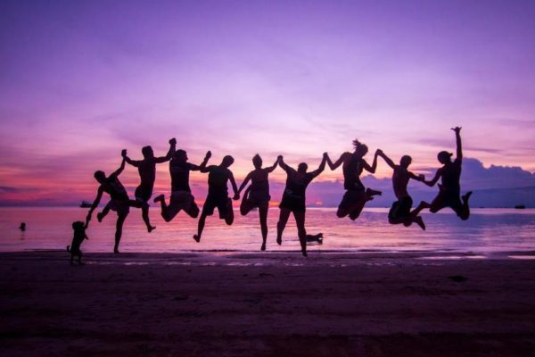 A group of people and a dog are silhouetted against a purple sunset sky, joyfully jumping together on a sandy beach, with a calm sea behind them.