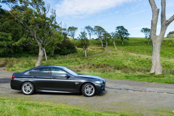A black sedan is parked on the roadside in a lush, green countryside setting with scattered trees and clear skies above.