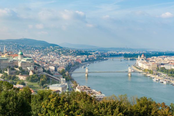 This is a panoramic view of a city bisected by a river, with bridges connecting the two sides, surrounded by greenery and hills under a cloudy sky.