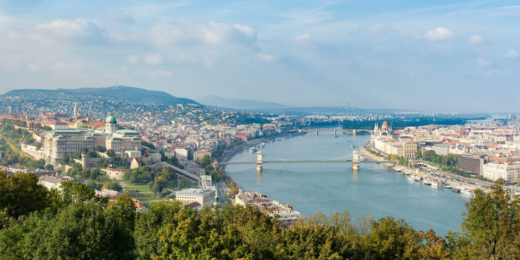 This is a panoramic view of a city bisected by a river, with bridges connecting the two sides, surrounded by greenery and hills under a cloudy sky.