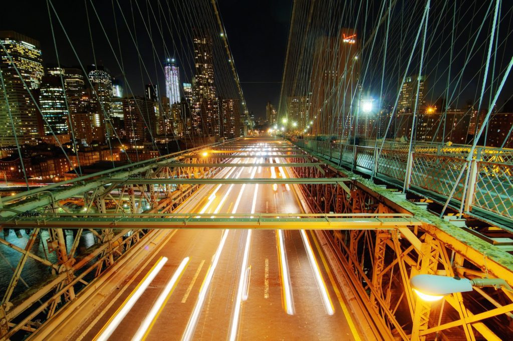 A night-time long-exposure photo capturing light trails on a busy bridge with steel cables, overlooking a city skyline illuminated against the dark sky.
