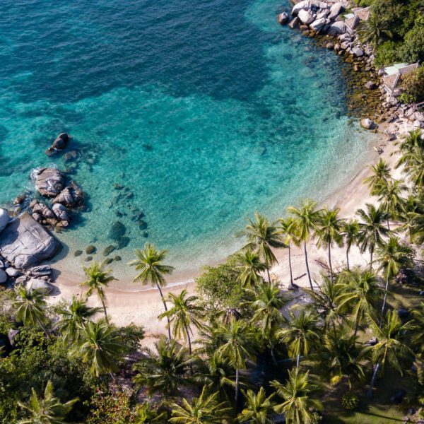 Aerial view of a tropical beach with clear turquoise water, white sand, lush palm trees, and large rocks dotting the serene shoreline.