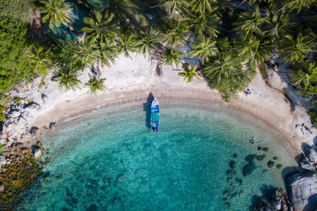 Aerial view of a tropical cove with crystal clear water, a boat in the center, surrounded by lush palm trees and a few people on the shore.