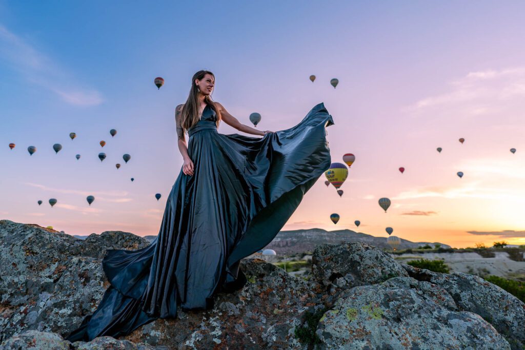 A person stands atop a rock at sunset, wearing a flowing black dress, with numerous hot air balloons in the sky behind them.