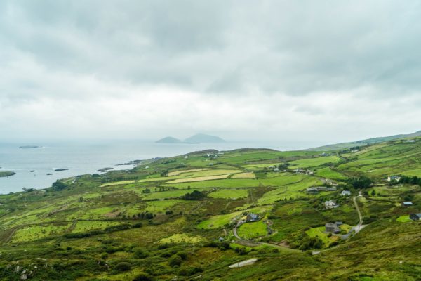 A serene, lush landscape with patchwork fields, winding roads, sporadic houses, surrounding waters, and cloud-covered mountains in the background.