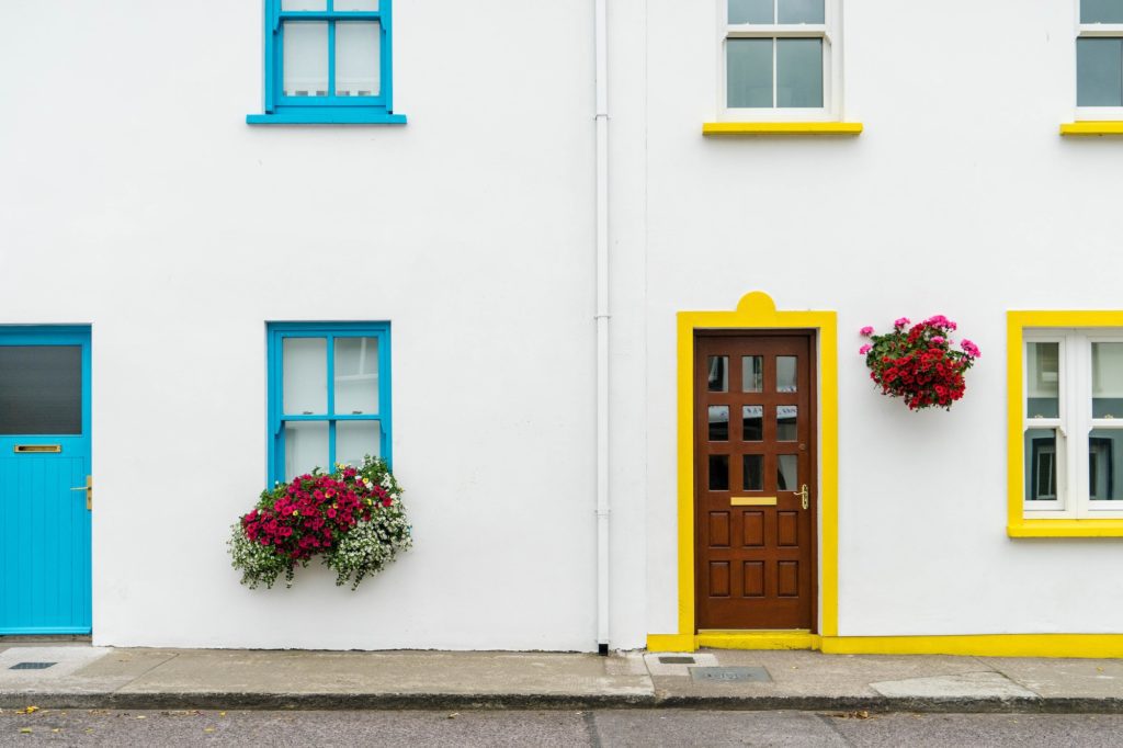 A colorful urban scene featuring two adjacent houses with brightly painted doors and windows, one blue, the other yellow, each adorned with a hanging flower basket.
