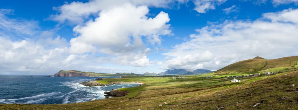 This panoramic image shows a scenic coastal landscape with rolling green hills, a blue sky with clouds, and a rugged coastline with cliffs.