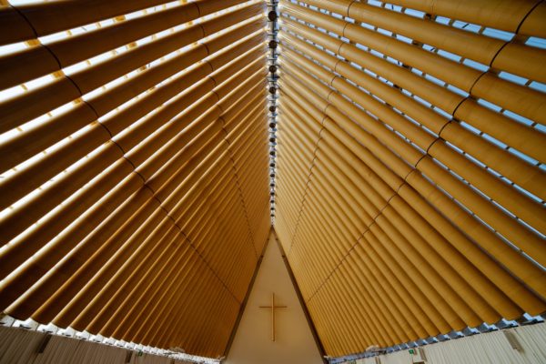 This image shows the interior ceiling of a modern church with symmetrical wooden beams converging towards a cross at the apex under a clear blue sky.