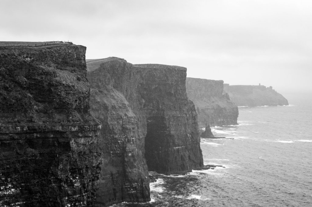 This black and white image shows towering, rugged cliffs overlooking a misty sea. The cliffs' edges are sharp against the overcast sky.