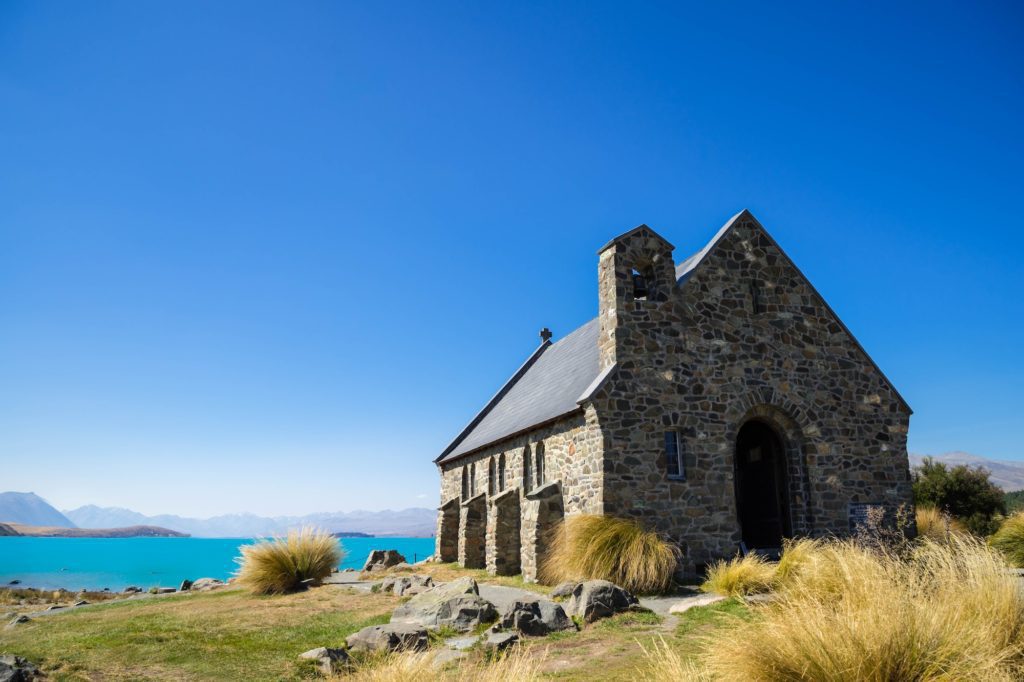 A stone chapel with a dark roof stands under a clear blue sky, surrounded by wild grass with a backdrop of a tranquil blue lake and mountains.