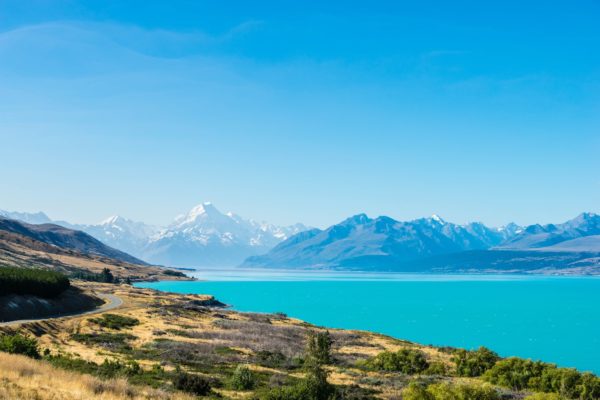 A stunning landscape showcasing a vibrant blue lake with snowy mountain peaks in the distance, clear skies, and a winding road alongside grassland.