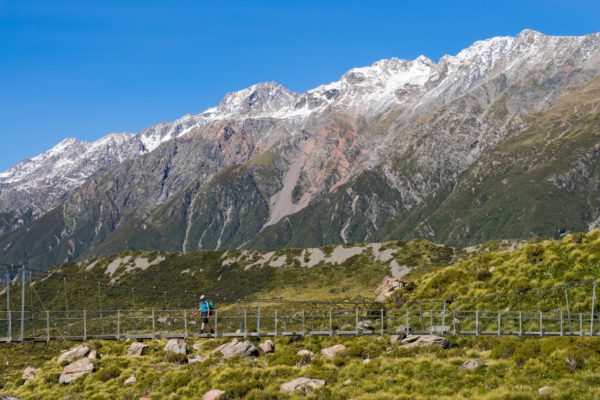 A person stands on a bridge amidst a scenic mountain landscape. Snow-capped peaks rise under a clear blue sky, with lush greenery surrounding the area.