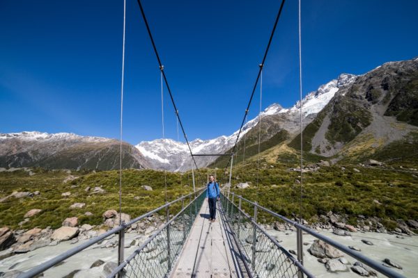 A person stands on a suspension bridge surrounded by mountains under a clear blue sky. Snow-covered peaks and rocky terrain stretch into the distance.