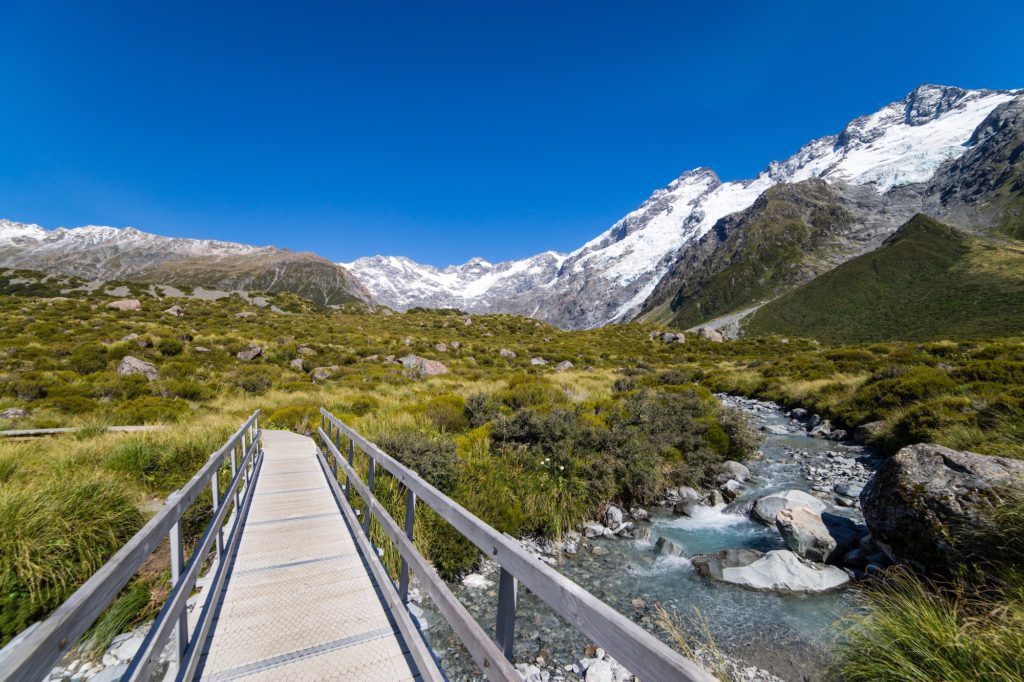 A metal footbridge spans a clear mountain stream, leading towards snowy peaks and alpine vegetation under a bright blue sky. A picturesque, tranquil natural scene.