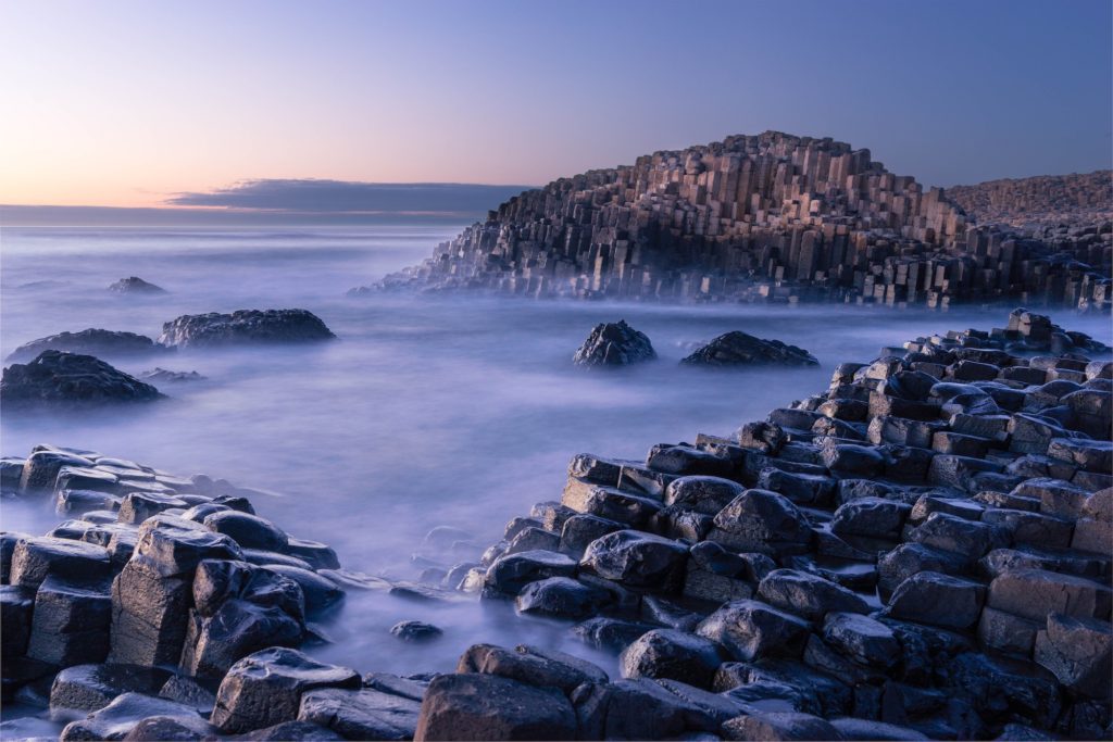 This is an image of the Giant's Causeway in Northern Ireland, featuring hexagonal basalt columns at the coast with misty waters and a soft-hued sky at dusk.