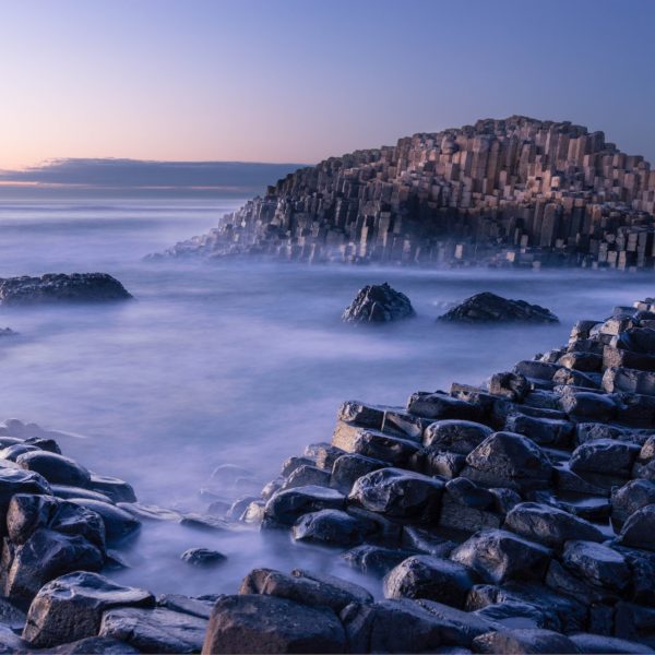 This is an image of the Giant's Causeway in Northern Ireland, featuring hexagonal basalt columns at the coast with misty waters and a soft-hued sky at dusk.