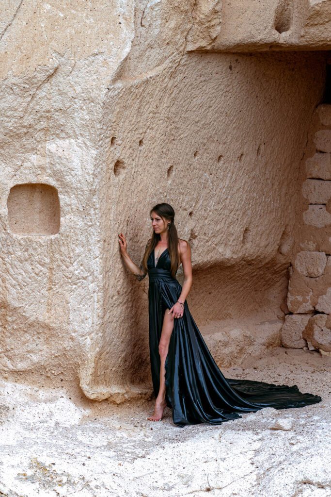 A person in a long black dress stands within an alcove in a sandy rock wall, touching the surface as fabric drapes elegantly to the ground.