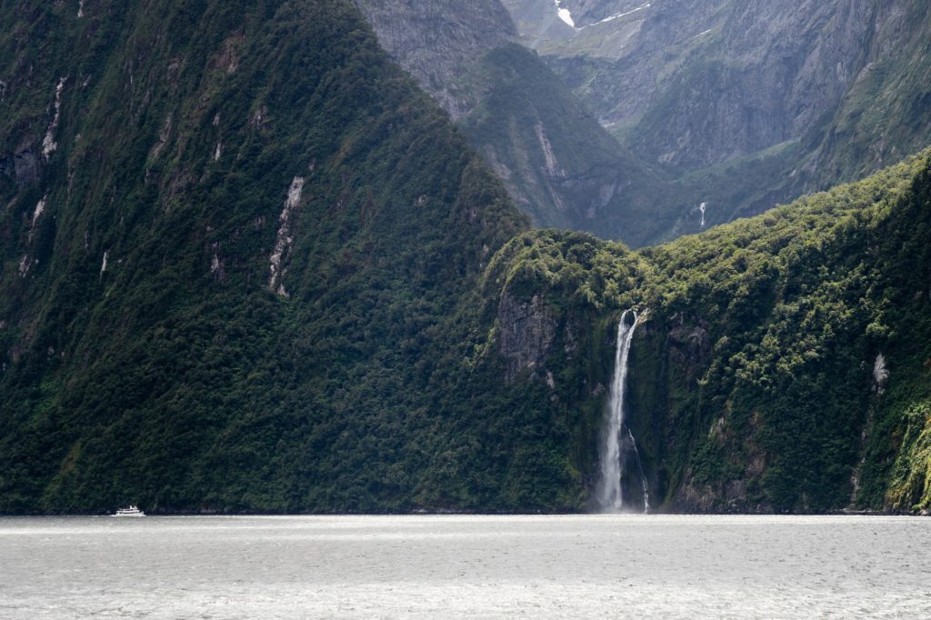 A serene landscape featuring a tall, slender waterfall cascading down a lush, green mountainside into a calm body of water, possibly a fjord or lake.