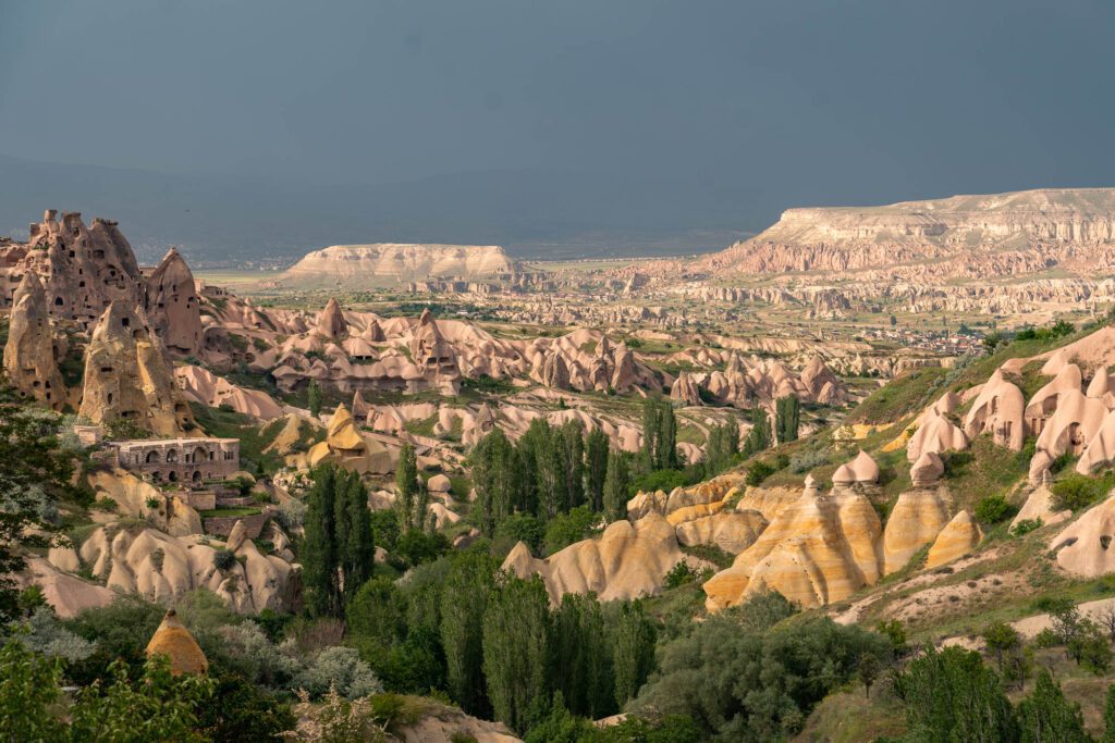A panoramic view of Cappadocia, Turkey, showcasing unusual rock formations, ancient cliff dwellings, green trees, and a dramatic, cloudy sky.