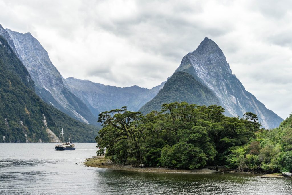A serene fjord landscape with a sailing boat near forested shores against a backdrop of dramatic mountain peaks under an overcast sky.