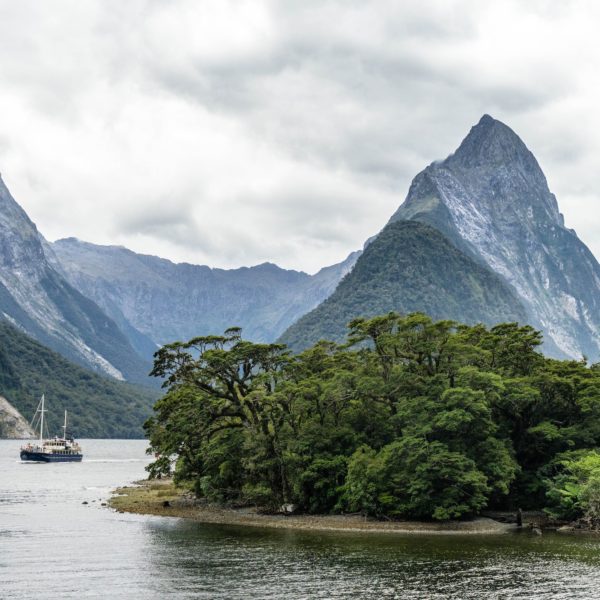 A serene fjord landscape with a sailing boat near forested shores against a backdrop of dramatic mountain peaks under an overcast sky.