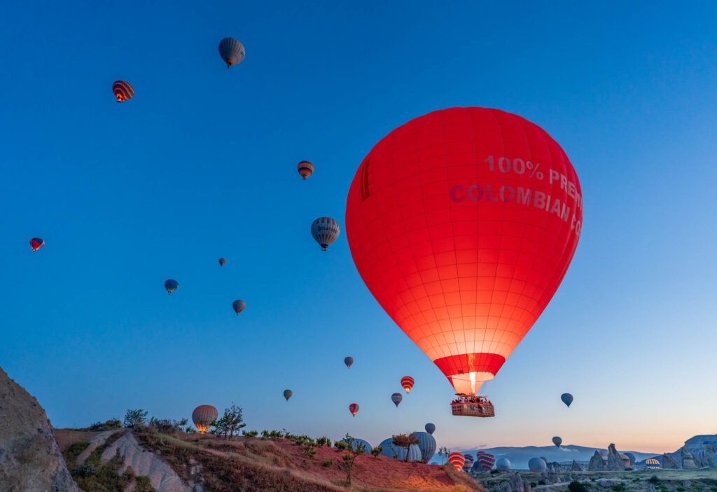 Numerous hot air balloons float in the sky at dawn, with a prominent red one closer to the viewer, over a rugged landscape.