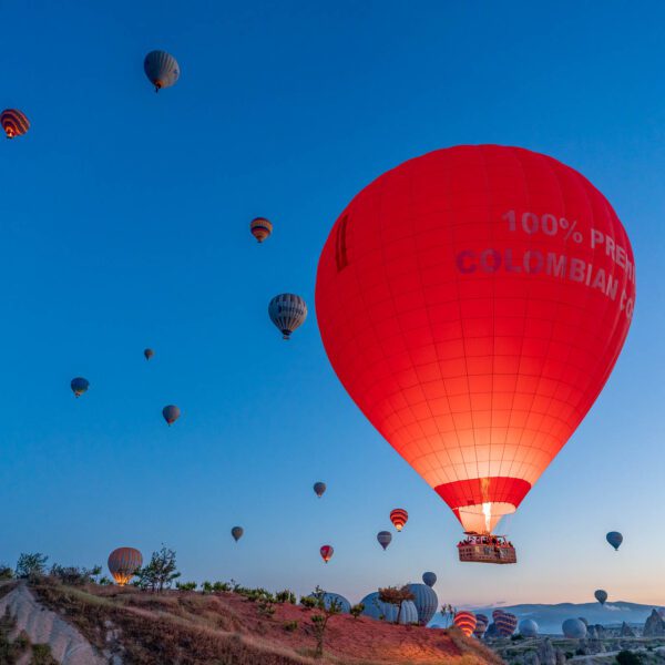 Numerous hot air balloons float in the sky at dawn, with a prominent red one closer to the viewer, over a rugged landscape.