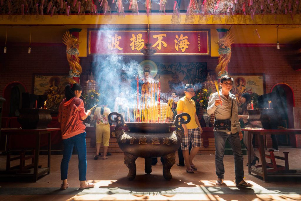 Inside a Chinese temple, people are burning incense sticks in a large urn. Sunlight streams through the smoke, illuminating traditional decorations and statues.