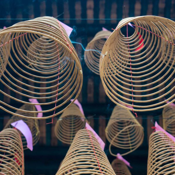 Circular incense coils hang from a ceiling, their delicate frames casting shadows below. Pink tags are attached to some, amidst a backdrop of wooden beams.